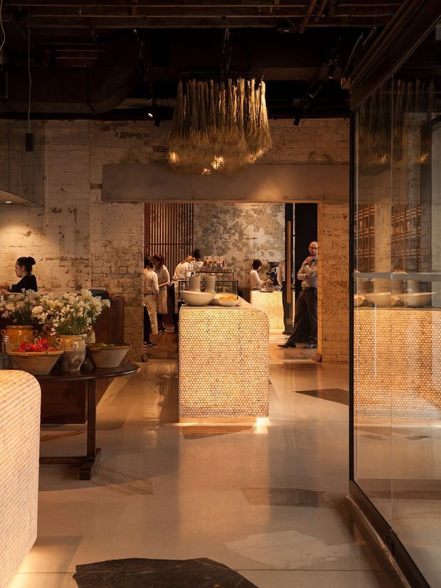 A warmly lit hospitality interior, likely a restaurant or bar, with exposed dark ceiling services, distressed pale brick walls, polished stone flooring, and a central rounded service island clad in small circular tiles. A dramatic suspended textured pendant hangs above the island, while glass partitions reflect the room and reveal additional bar or display joinery. Staff and dining activity are visible in the background, with floral arrangements on a dark timber table in the foreground.