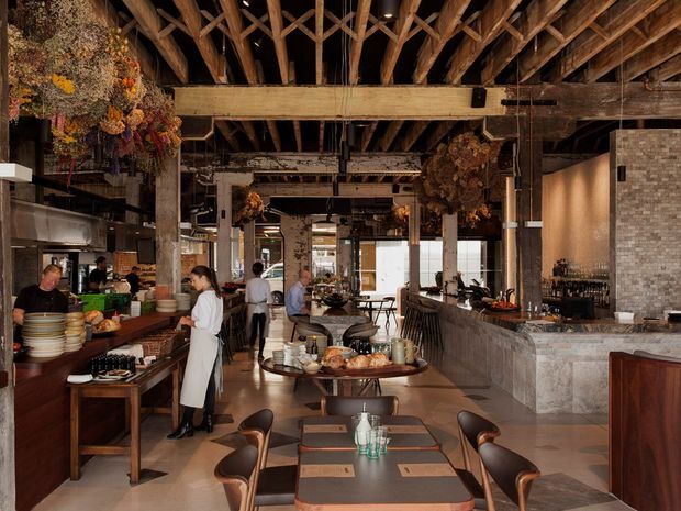 A spacious, atmospheric restaurant interior with an open kitchen and bar area, communal and individual dining tables, upholstered dining chairs, and extensive exposed timber roof trusses and beams. The space combines dark timber, textured masonry, concrete-toned surfaces, hanging dried floral arrangements, and warm ambient lighting in an industrial, rustic setting.