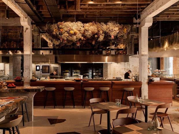 A large industrial-style restaurant or commercial dining space with an open kitchen and long timber-fronted counter. Exposed timber ceiling structure, rough painted masonry columns and walls, stainless-steel kitchen equipment, and dark metal details create a warm, rustic-industrial atmosphere. Bar stools line the counter, while small timber dining tables and chairs occupy the foreground. A dramatic suspended arrangement of dried flowers and foliage hangs above the kitchen counter.