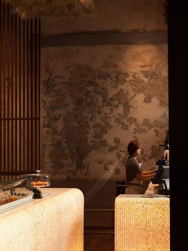 Warm, moody interior photograph of a contemporary café or hospitality counter. The scene features two rounded counters with illuminated honeycomb-patterned fronts, a dark timber vertical-slat screen, a heavily textured plaster feature wall, timber flooring, and a barista working behind the service counter. Low amber lighting creates an intimate, atmospheric setting.