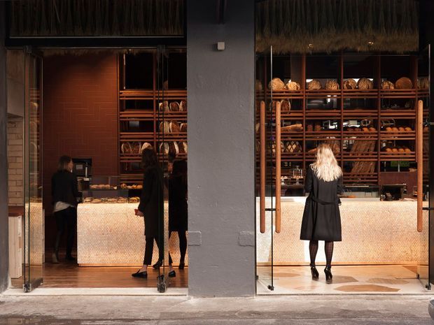 Front-facing view into a warm, contemporary bakery or bread shop through large glazed openings. Dark timber shelving is densely stocked with loaves and baked goods, while illuminated pale mosaic-faced service counters sit inside. Rich terracotta-toned wall tiles, timber flooring, and a central rendered structural pier frame the commercial interior. Customers and staff are visible, with the street threshold and exterior pavement in the foreground.