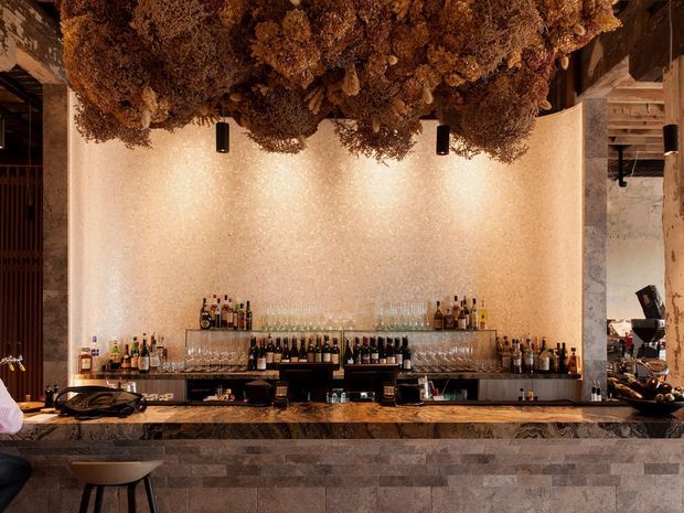 A warmly lit commercial bar interior with a long stone-faced counter, dark timber or metal bar shelving, extensive glassware and liquor displays, and a textured illuminated wall behind the bar. A dense dried botanical installation hangs across the ceiling, adding a dramatic natural feature. Partial stools and service equipment are visible at the edges.