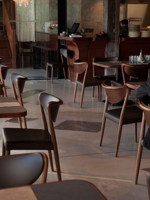 A refined, dimly lit restaurant interior with numerous dark timber dining chairs and tables arranged across a polished stone floor. A curved timber reception or service counter occupies the rear, backed by textured aged stone or plaster wall panels and dark structural columns. Glass partitions, shelving with stacked crockery, and warm decorative lighting contribute to an upscale, moody hospitality atmosphere.