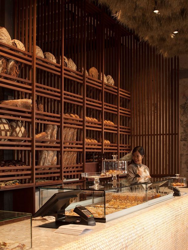 Warmly lit commercial bakery or pastry shop interior with a long display counter in the foreground and extensive dark timber shelving behind it, stocked with breads, pastries, and baskets. A staff member stands behind the counter. The space features richly toned vertical timber slats, glass display cases, a textured light-coloured counter front, and a dramatic shaggy suspended ceiling feature.