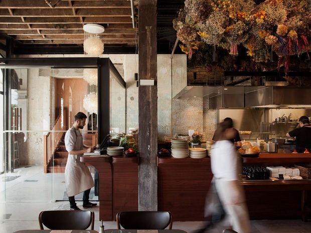 A warmly lit restaurant interior with an open commercial kitchen and dining area. The space features exposed timber ceiling joists, substantial dark timber structural posts, glass partitions, dark wood cabinetry and counters, stainless-steel kitchen equipment, and a dramatic installation of dried floral arrangements suspended overhead. Staff are working behind the counter, while dining furniture is visible in the foreground.