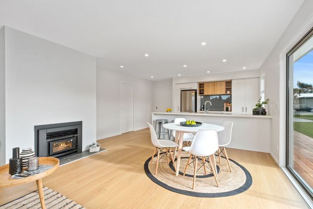 Bright open-plan kitchen and dining area with white cabinetry, a pale stone island bench, timber flooring, a round white dining table with modern chairs, and a built-in fireplace. Large sliding glass doors open to an outdoor deck, creating an airy contemporary interior.