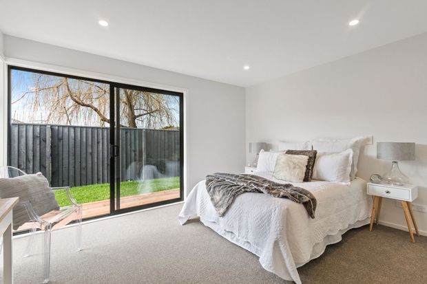 A bright, minimalist bedroom with a white upholstered bed dressed in layered white bedding, textured throws, and neutral cushions. A compact timber bedside table and lamp sit beside the bed, while full-height black-framed sliding glass doors open visually to a small lawn and fenced outdoor area. The room has recessed ceiling lights, pale walls, and soft beige carpet.