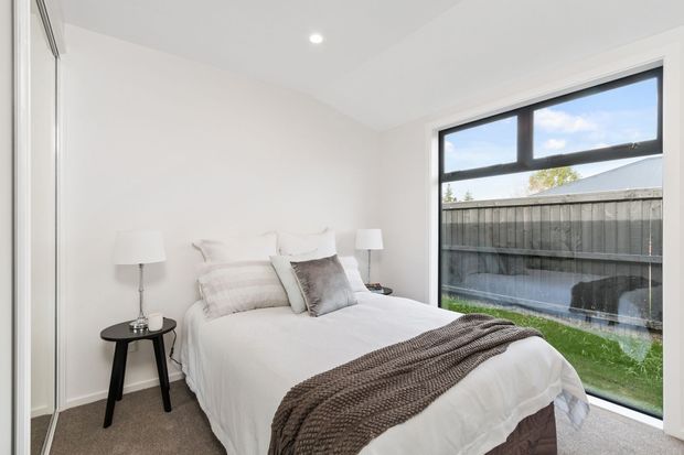 Bright, compact contemporary bedroom with a neatly made bed, white bedding, layered neutral pillows, a textured taupe throw, black side table, bedside lamps, and large black-framed window overlooking a fenced outdoor area. The room has white walls and ceiling, carpeted flooring, and a clean, understated fit-out.