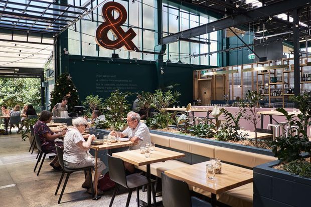 A bright contemporary commercial dining venue with an open industrial-style ceiling, extensive floor-to-ceiling glazing, exposed dark steel beams and services, timber dining tables, upholstered chairs, and large integrated planter beds dividing seating areas. A prominent illuminated ampersand sign and a bar or service counter are visible in the background.