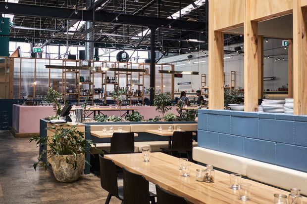 A spacious contemporary restaurant interior with an open dining area, long timber tables, dark upholstered chairs, built-in cream banquette seating, and a blue painted block partition. Timber-framed service and display structures, indoor plants, exposed black ceiling services, and extensive glazing create an industrial hospitality setting with warm natural accents.