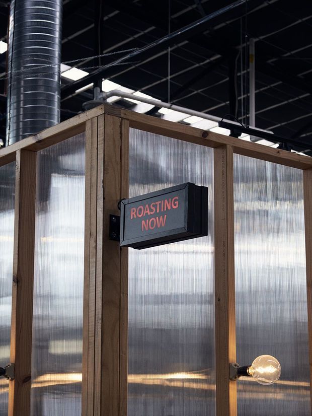 Close-up of an industrial commercial interior enclosure framed in timber and clad with translucent corrugated panels. A black illuminated sign reads “ROASTING NOW,” with exposed ductwork, dark ceiling services, and a visible industrial-style light fitting.