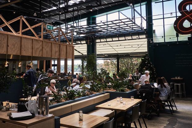 A spacious industrial-style restaurant or café interior with a high exposed ceiling, extensive floor-to-ceiling glazing, black metal framing, timber-framed mezzanine or service structure, and abundant indoor planting. Dining tables, banquette seating, bar stools, pendant lights, and patrons fill the open space.