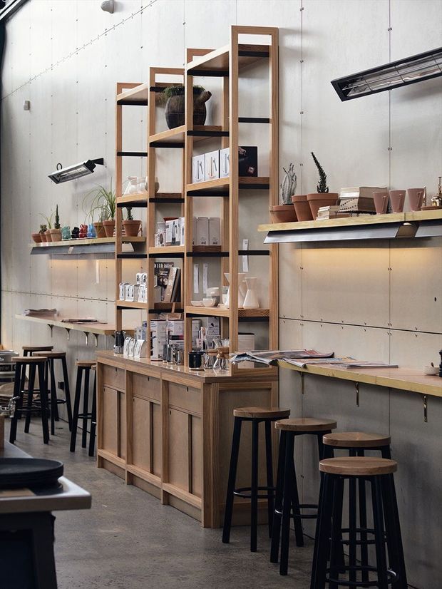 A compact, industrial-style café or retail showroom interior with exposed pale concrete walls, timber shelving and cabinetry, long built-in counters, black metal stools, display merchandise, potted plants, and linear wall-mounted lighting. The space combines warm wood joinery with a cool grey architectural shell and appears designed for browsing, sitting, and product display.