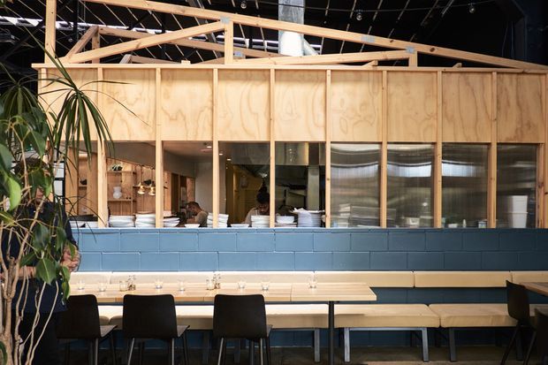 Interior view of a contemporary commercial dining or hospitality space, with a long communal dining table and dark chairs in the foreground. A blue-painted masonry half-wall supports a timber-framed plywood partition and glazed service or kitchen area. Exposed timber roof trusses, open ceiling services, pendant lights, and indoor planting contribute to an industrial and warm utilitarian character.