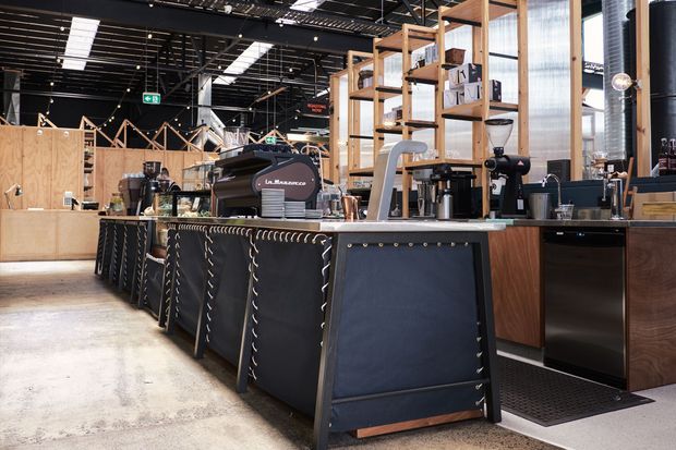 A spacious industrial-style commercial café interior with a long coffee service counter, espresso machines, grinders, timber shelving, and black leather-like panel detailing. The space features exposed dark ceiling structure, polished concrete flooring, timber screens, and large glazed openings.