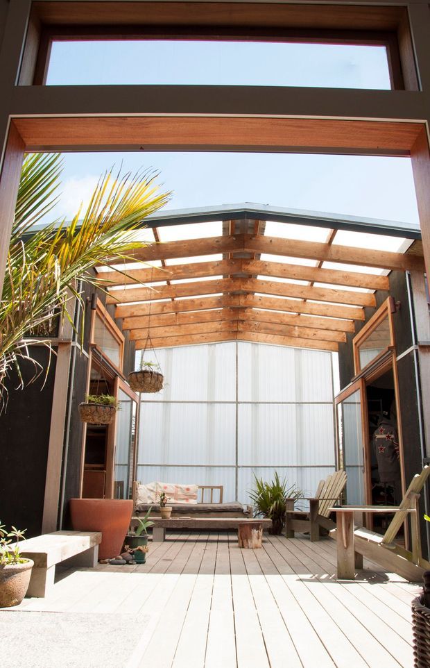 A bright enclosed courtyard or sunroom with a timber-decked floor, large timber-framed openings, and a partially open timber pergola roof supported by dark structural posts. The space contains built-in and freestanding timber seating, potted plants, hanging planters, and a small central stump table. Translucent wall panels provide privacy and abundant daylight, creating an airy indoor-outdoor living area.