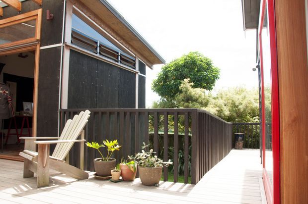 A bright exterior view of a timber-clad elevated deck and covered walkway beside a dark-clad building. The deck has dark vertical balustrades, pale timber decking, a red-framed glazed wall, an open doorway, an Adirondack-style chair, and potted plants. A gently sloping ramp or walkway leads toward landscaped surroundings, with exposed timber framing visible overhead.