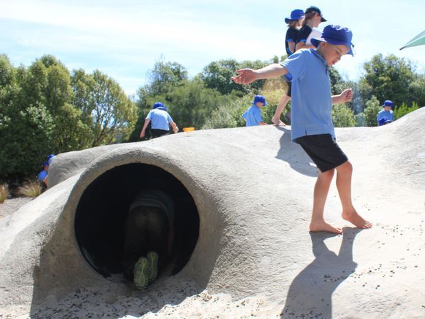 Outdoor playground scene featuring children climbing and crawling over a large sculptural, concrete-like play structure with a dark tunnel opening. Trees and open sky form the background.