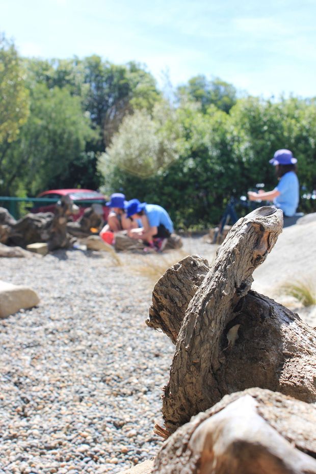 A bright outdoor landscaped play area with a broad pebble-covered ground surface, large natural weathered logs in the foreground and children playing with logs and rocks in the background. Dense green planting surrounds the space, creating an informal nature-play setting.