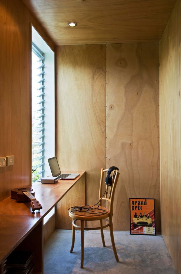 Compact wood-lined workspace with plywood walls and ceiling, a built-in timber desk beneath a louvred window, laptop, guitar, headphones, and a simple wooden chair. The room has a warm, intimate studio-like atmosphere with a polished concrete floor and recessed ceiling light.