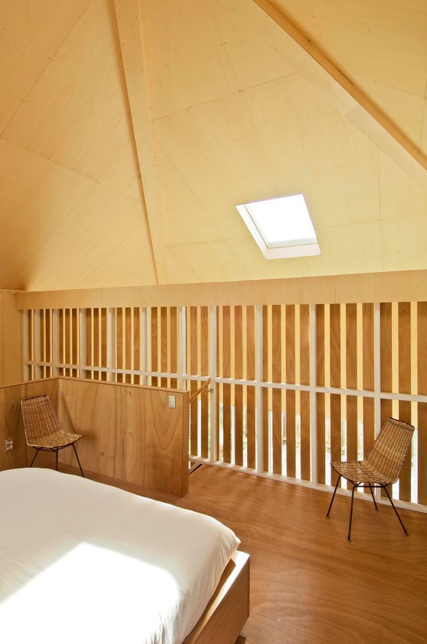 A warm, wood-lined bedroom in a loft-like upper-level space. The room features a pitched timber ceiling with exposed structural beams, a rectangular skylight, timber flooring and wall panelling, and a white-painted vertical balustrade overlooking the level below. A timber bed with white bedding and two woven chairs complete the simple interior.