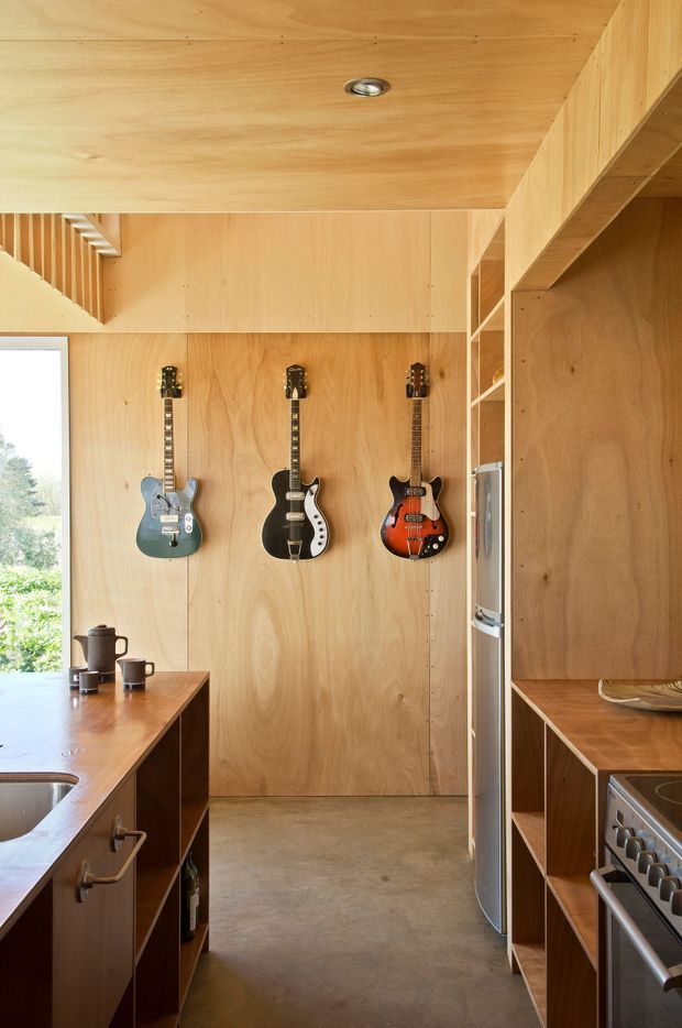 Compact kitchen with extensive warm plywood wall, ceiling, island and open shelving joinery. A refrigerator and stainless steel range sit along the right side, while the island includes a sink and timber benchtop. Three guitars are mounted on the rear plywood wall, adding a distinctive creative accent. A large window brings in daylight and views of greenery.