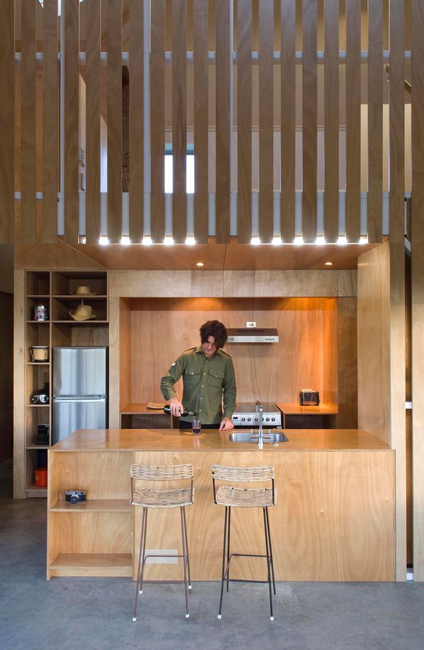 A compact, vertically oriented kitchen with extensive warm timber veneer joinery. A central island with integrated sink and two woven bar stools faces a cooktop and rangehood set into a timber-lined backsplash. Open shelving and a stainless-steel refrigerator sit to the left, while a dramatic upper screen of closely spaced vertical timber battens filters light beneath a high ceiling. The space has a clean contemporary character with warm, natural materials and concrete flooring.