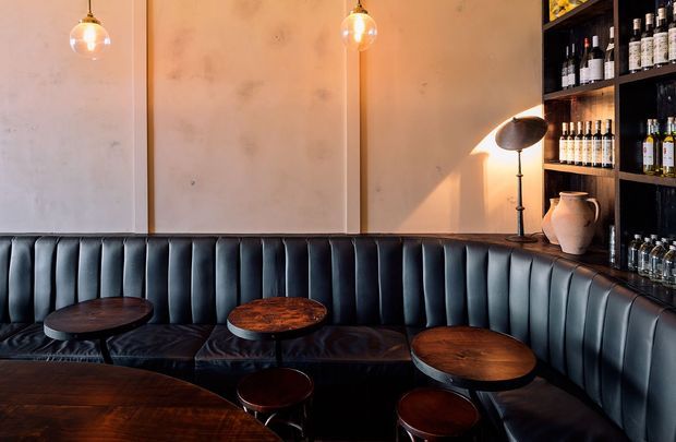 A moody, sophisticated bar or lounge interior with a curved, channel-tufted dark leather banquette wrapping around small round wooden tables. Warm pendant lights, a floor lamp, plaster walls, and dark built-in shelving displaying bottles create an intimate hospitality setting.
