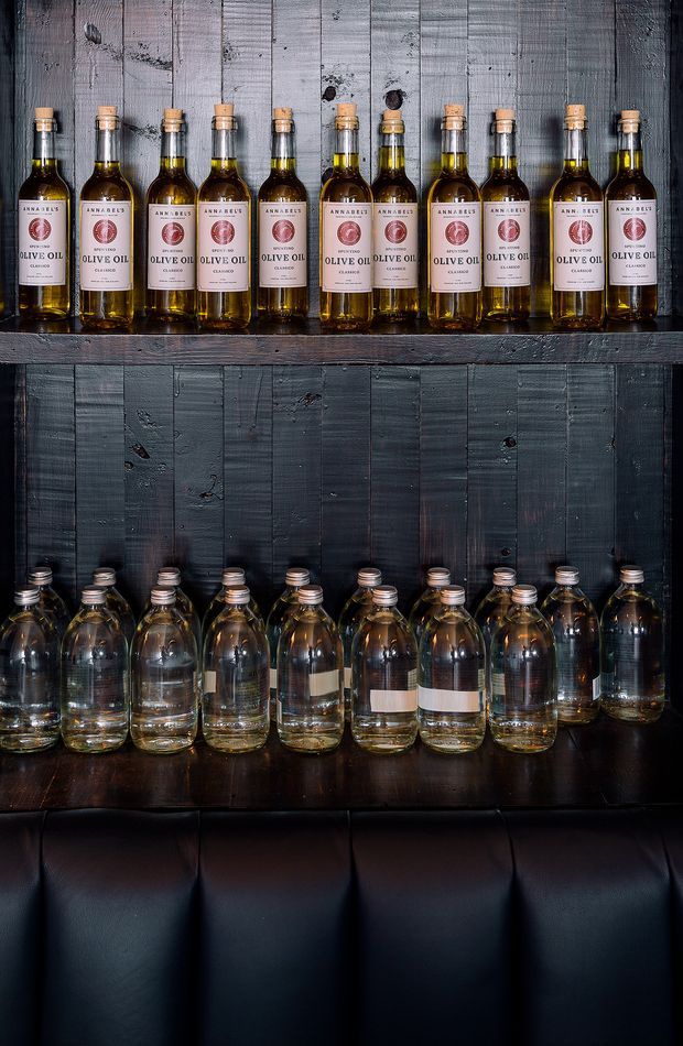 A dark, moody product display featuring rows of olive oil bottles arranged on rustic black-painted timber shelves against a distressed timber wall. The lower shelf holds larger glass bottles, and a black leather-upholstered surface appears below.