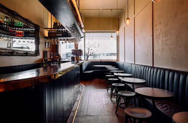 A narrow, stylish bar and dining lounge with a long dark timber bar counter on the left, mirrored bottle shelving, black vertical-panel bar front, and a continuous dark banquette along the right wall. Small round tables and stools line the seating area, while warm pendant lights hang from a pale tiled ceiling. Large windows at the rear provide bright daylight and an urban outlook.