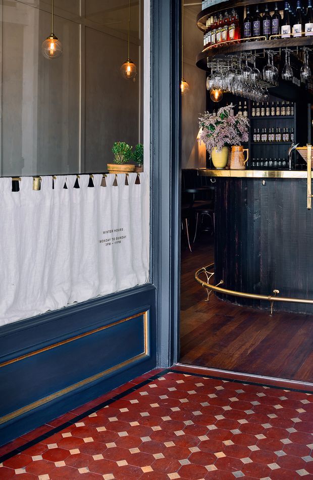 View through the entrance of a refined, dark-toned bar or hospitality venue. The foreground features a richly patterned red and cream geometric tiled floor and a deep navy-blue door with inset gold trim. Inside, a black bar with brass edging, dark timber flooring, illuminated bottle shelving, hanging glassware, pendant lights, stools, and floral arrangements create an intimate, vintage-inspired atmosphere.