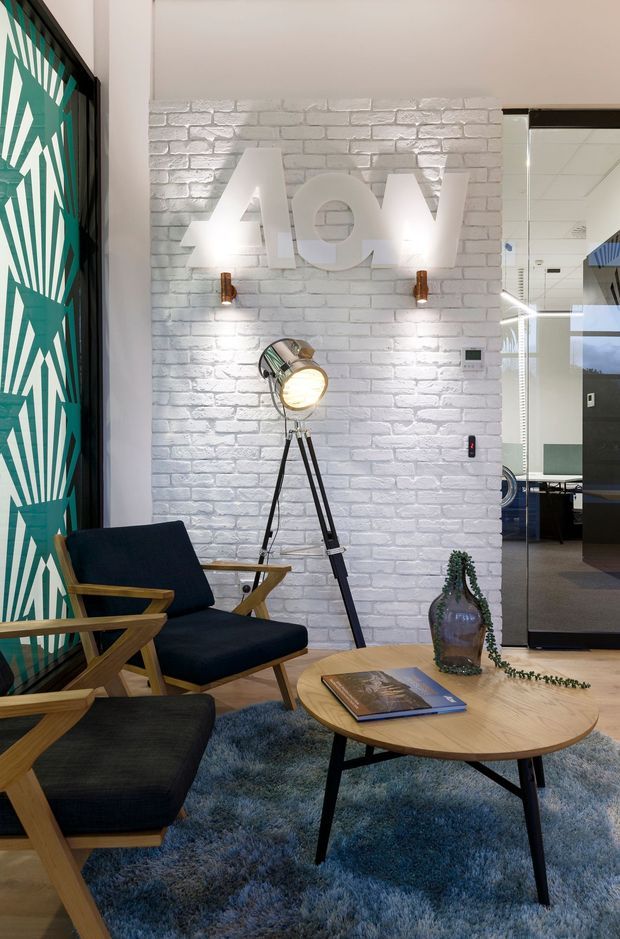 A stylish commercial reception or lounge area with a white painted brick feature wall, illuminated raised lettering, and warm timber-and-black furnishings. The space includes a tripod floor lamp, sculptural wall sconces, a patterned teal and white glazed partition or door, a round timber coffee table, dark upholstered lounge chairs, and a blue-grey shag rug. A glazed opening reveals an adjacent office area.