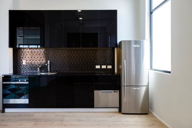 A compact contemporary kitchen with glossy black cabinetry, a large black splashback area featuring a patterned metallic tile backsplash, dark benchtops, stainless-steel refrigerator, integrated dishwasher, and a wide sunlit window. Pale timber flooring and white walls provide contrast.