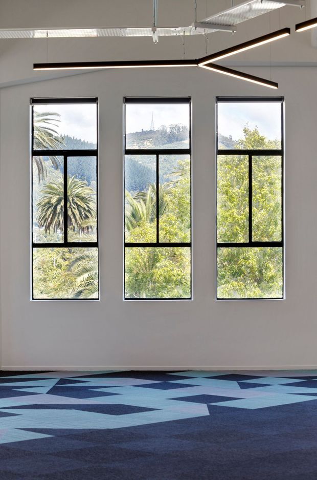 A bright, minimally furnished commercial interior with three tall black-framed windows overlooking lush palm trees and a hilly landscape. The space has white walls, exposed services and suspended fluorescent lighting, distinctive branching linear pendant lights, and a bold geometric blue patterned carpet.