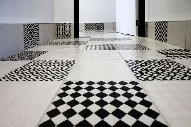 Low-angle view of a tiled interior floor featuring a bold patchwork of black-and-white geometric tile patterns, including checkerboard, diamonds, lattice, and repeating motifs. Matching patterned tile sections continue up the lower walls, with white walls and a doorway in the background.