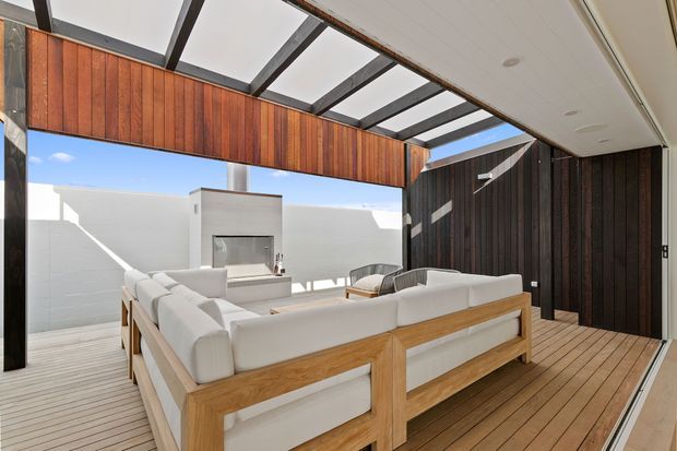 Bright contemporary outdoor lounge beneath a partially glazed pergola, featuring a large white sectional sofa with timber framing, timber decking, dark vertical timber wall cladding, and a built-in outdoor fireplace or barbecue against a white masonry wall. Strong indoor-outdoor connection and exposed structural beams define the space.