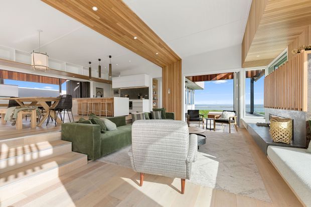 Bright open-plan coastal interior with a living room in the foreground, dining area and kitchen beyond, and large glazed openings framing a visible ocean view. The space features extensive warm timber ceiling and wall lining, pale timber flooring, a green sofa, woven and upholstered seating, a stone fireplace or feature wall, and a restrained contemporary fit-out. Split-level steps connect the living and dining zones.