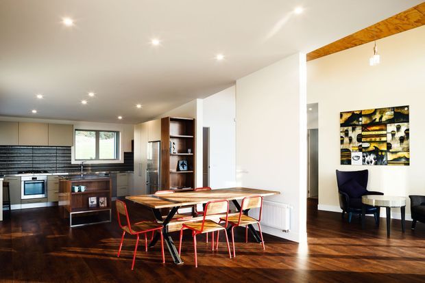 Open-plan interior combining a contemporary kitchen, dining area, and adjacent sitting space. The room features dark timber flooring, white walls and ceiling, dark cabinetry, a timber dining table with bright red metal-framed chairs, recessed ceiling lighting, and a timber ceiling beam. A compact lounge area with an armchair and artwork is visible beyond the dining space.