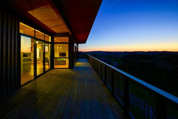 A wide timber deck or balcony extends along a modern building at dusk, with dark vertical railings, expansive glazing, warm interior lighting, and a broad view over a wooded landscape and sunset horizon. The deep roof overhang and timber-lined soffit create a sheltered outdoor transition space.