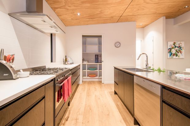 A bright contemporary kitchen with a central island, extensive dark timber cabinetry, light timber flooring, white benchtops, stainless-steel appliances, and a warm plywood-lined ceiling. The room includes a large cooking zone on the left, a sink and dishwasher integrated into the island, open shelving at the rear, recessed lighting, and a small floral artwork.