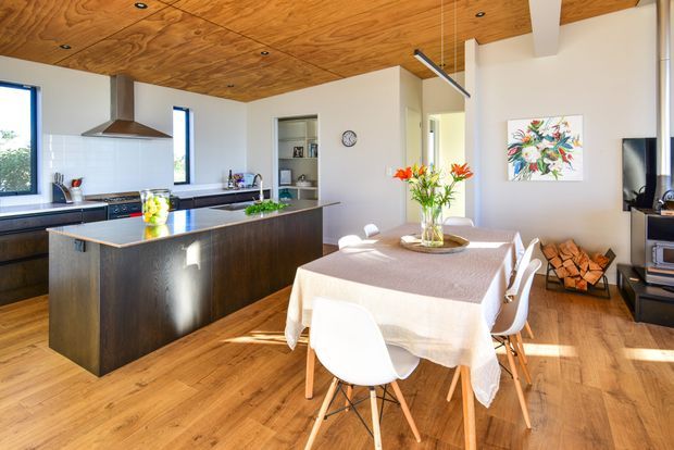 Bright open-plan kitchen and dining area with a large dark-wood island, stainless-steel appliances, white cabinetry, timber flooring, and a distinctive exposed timber ceiling with recessed spotlights. A contemporary dining table and white chairs occupy the foreground, while a living area with a television and fireplace is visible to the right.
