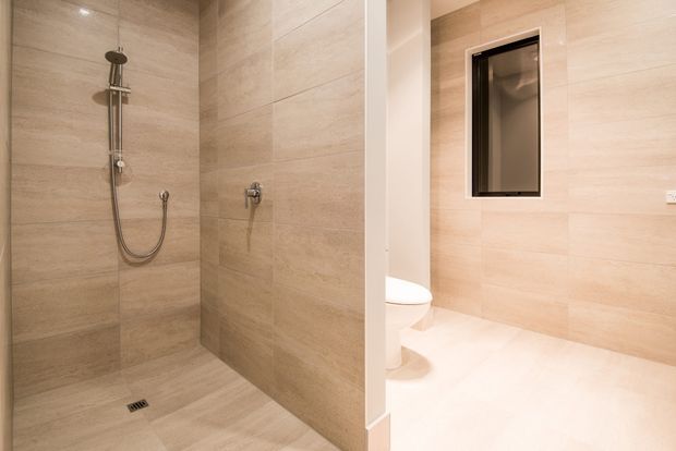 A contemporary bathroom featuring a walk-in tiled shower with a wall-mounted rain shower and handheld fixture, a concealed toilet area, and a small high-set window. The room is finished throughout in warm beige stone-look tiles with minimal chrome fittings and bright recessed lighting.