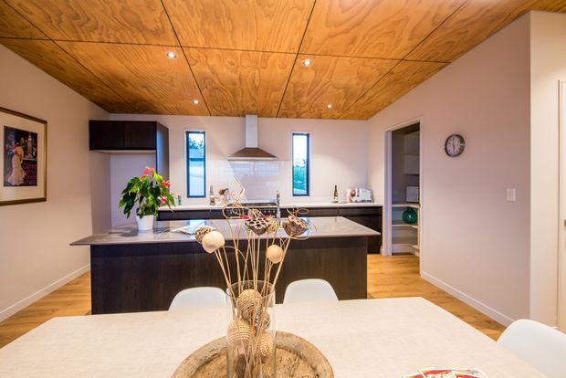 Open-plan kitchen and dining area with a dark timber island, pale stone benchtops, integrated appliances, narrow vertical windows, and a striking plywood-lined ceiling with recessed downlights. A white dining table and sculptural floral arrangement occupy the foreground, while warm timber flooring and neutral walls create a contemporary, warm-toned interior.