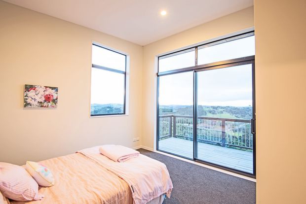 A bright, compact bedroom with a bed, soft pink bedding, beige walls, carpeted flooring, and large black-framed sliding glass doors opening onto a balcony with expansive landscape views. A small artwork hangs on the wall, and a recessed ceiling light provides illumination.