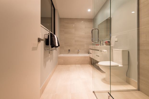 Contemporary bathroom with a freestanding-style bathtub at the rear, glass-enclosed walk-in shower, wall-mounted vanity, toilet, heated towel rail, and large-format beige stone-look wall and floor finishes. The space has warm neutral tones, clean lines, and recessed ceiling lighting.