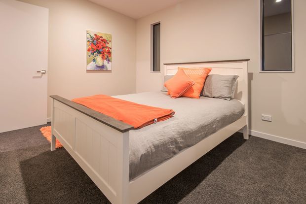 A compact, warmly lit bedroom with a white panelled bed featuring a dark timber-look top rail, grey bedding, and bright orange cushions and throw. The room has beige painted walls, dark grey carpet, a narrow vertical window, a dark-framed mirrored or glazed wardrobe panel, recessed ceiling lighting, and colourful wall artwork.