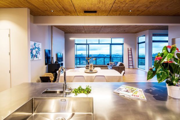 Open-plan contemporary kitchen overlooking a bright living and dining area, with a large stainless-steel island sink and benchtop in the foreground. The room features extensive floor-to-ceiling glazing, warm timber ceiling lining, pale walls, timber flooring, and views toward a blue waterfront or coastal landscape. A dining table, sofa, television, artwork, and indoor plants are visible beyond the kitchen.