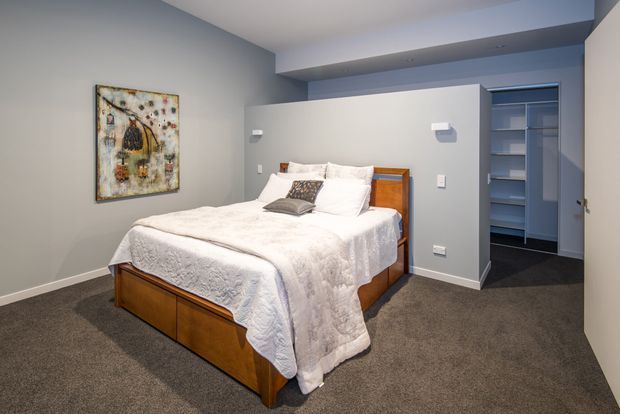 A compact modern bedroom with a wooden bed and white bedding, set against soft grey walls and dark grey carpet. A concealed or partially enclosed walk-in wardrobe with built-in shelving is visible behind the bed, while recessed ceiling lighting and simple wall-mounted fixtures create a clean, contemporary feel.