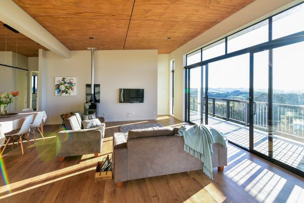 Bright contemporary open-plan living and dining area with warm timber flooring and a timber-lined ceiling. The room features large black-framed floor-to-ceiling glazing opening to a balcony, with expansive elevated views beyond. A sofa arrangement faces a wall-mounted television, while a dining table and chairs sit near a mirrored wall.