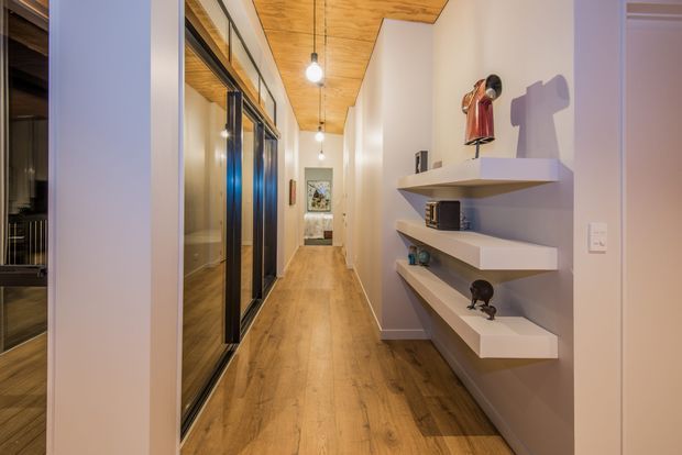Long, narrow contemporary hallway with warm timber flooring and a matching timber-lined ceiling. Full-height dark-framed glazed doors or partitions line the left side, while white built-in floating display shelves with small decorative objects occupy the right wall. Simple pendant lights and light neutral walls create a warm, refined circulation space.